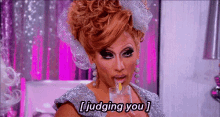 a drag queen is drinking from a glass and saying " judging you . "