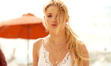 a woman in a white tank top is standing in front of an orange umbrella on the beach .
