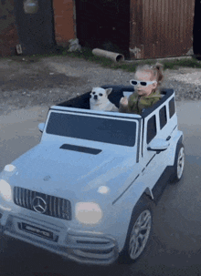 a little girl is driving a white mercedes with a dog in the back seat