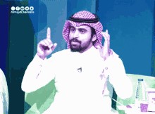 a man wearing a head scarf and a white shirt is sitting in front of a sign that says almajdchannels