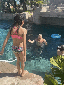 a little girl in a pink bikini stands on the edge of a swimming pool