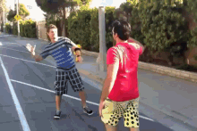 a man in a red shirt is standing next to a man in plaid shorts on a street