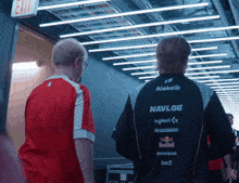 two men are walking down a hallway and one of them is wearing an aleksib navi.gg jacket