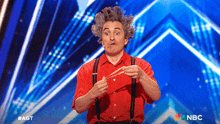 a man in a red shirt and suspenders performs on a stage with a nbc logo in the background