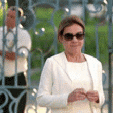 a woman wearing sunglasses and a white jacket is standing in front of a gate