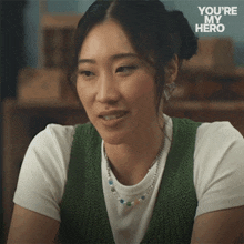 a woman wearing a green vest and a white shirt with the words you 're my hero behind her