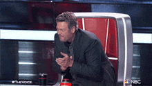 a man in a suit sits in a chair with a nbc logo in the background