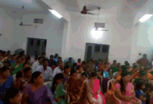 a large group of people sitting in a room with a ceiling fan
