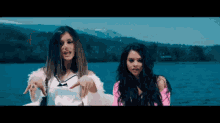 two women are standing next to each other in front of a body of water with mountains in the background