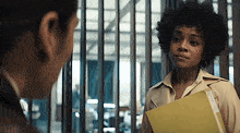 a woman in a trench coat is holding a yellow folder and talking to a man behind bars .