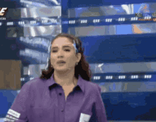 a woman wearing a purple shirt and a face shield is standing in front of a television screen .