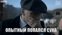 a man in a suit and hat is covering his face with his hand .