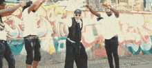 a group of people are dancing in front of a wall with graffiti on it that says this