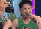 a man in a green tank top is singing into a microphone while another man looks on ..
