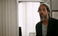 a man with a beard is standing in front of a window and looking at the camera .
