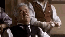 a man in a suit and tie is sitting in a chair while a man in a vest is standing behind him .