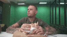 a man with tattoos is sitting at a table eating a large sandwich .