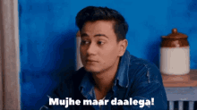 a man in a denim shirt is sitting in front of a blue wall and says mujhe maar daalega !