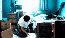 a man is kneeling on the floor in a messy room with boxes and a fan on the wall