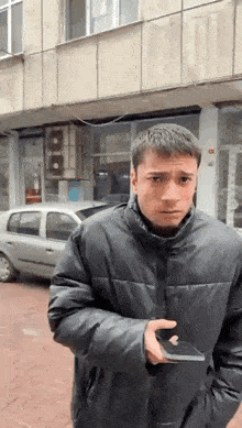 a man in a black jacket is holding a cell phone in front of a building and a car .