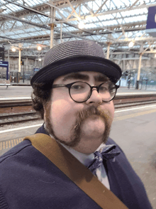 a man wearing a hat and glasses is standing in front of a sign that says ' railway station ' on it