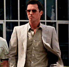 a man wearing sunglasses and a suit stands in front of a window
