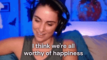 a woman wearing headphones is sitting on a bed and saying `` i think we 're all worthy of happiness ''