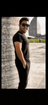 a man wearing sunglasses and a black t-shirt is standing in front of a building .