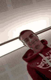 a man wearing glasses and a red hoodie is standing in front of a wall and smiling .