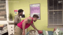 two men are standing next to each other in a bedroom . one of the men is wearing a pink tank top .