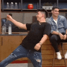 a man in a black shirt is dancing while another man sits on a bar stool
