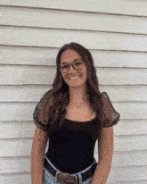 a woman wearing glasses and a black top is smiling in front of a white wall