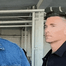 a man in a denim jacket and a black shirt is standing next to another man in a black shirt
