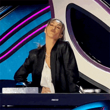 a woman in a black jacket is standing in front of a rbd3d television