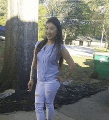 a woman in a blue shirt and white jeans stands in front of a tree