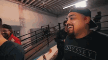 a man wearing a black odyssey t-shirt smiles in a room
