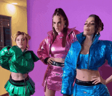 three women are standing in front of a purple wall wearing different colored outfits