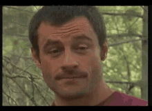 a man with a mustache is making a funny face in front of trees .