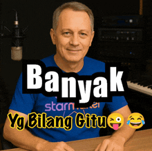 a man in a blue shirt is sitting in front of a microphone with the words banyak yg bilang citu behind him