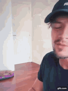a man wearing a hat and a black shirt is sitting in a room with a skateboard on the floor .