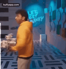 a man in a yellow hoodie is playing drums in a room with a sign that says let 's get funky .