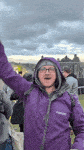 a man wearing a purple jacket and glasses is standing in a crowd with his arms in the air .