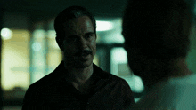a man with a mustache talks to another man in the dark
