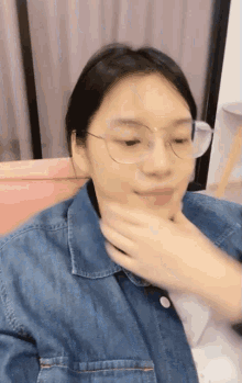 a woman wearing glasses and a denim jacket is covering her mouth with her hand