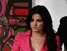a woman in a pink jacket and necklace is standing in front of a painting
