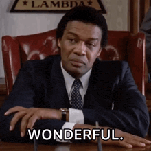 a man in a suit and tie sits at a desk with his hands on his hips and says wonderful
