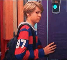 a young boy is standing in front of a purple locker with a backpack .