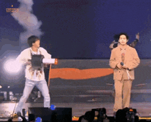 a man in a suit is holding a microphone while another man in a white shirt is holding a gun on stage