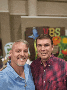 two men are posing for a picture in front of a vbs sign