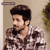 a man in a plaid shirt is sitting next to a wall and looking at the camera .
